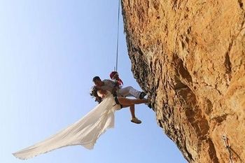 Rock Climbing wedding couple kissing while suspended on rope next to tan overhanging cliff.